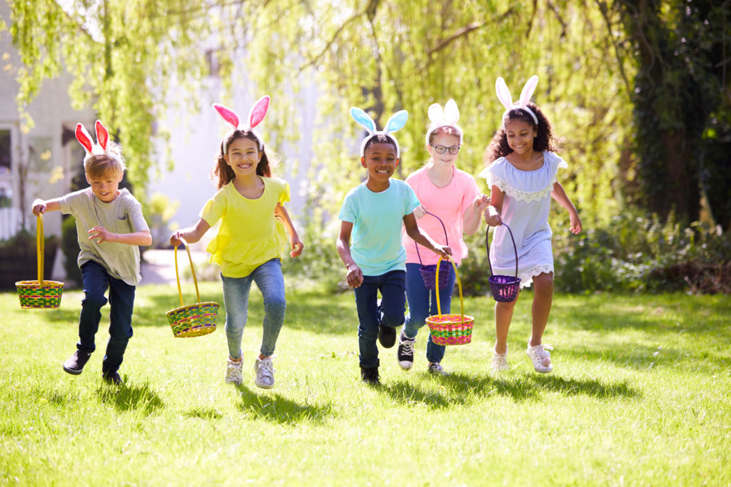 Group Of Children Wearing Bunny Ears Running To Pick Up Chocolate Egg On Easter Egg Hunt In Garden